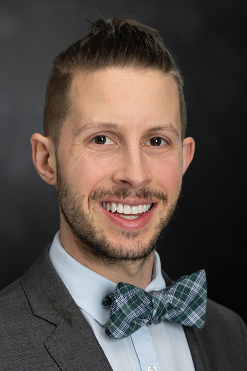 Man wearing a dark grey suit, light blue shirt, plaid bowtie grins at camera.